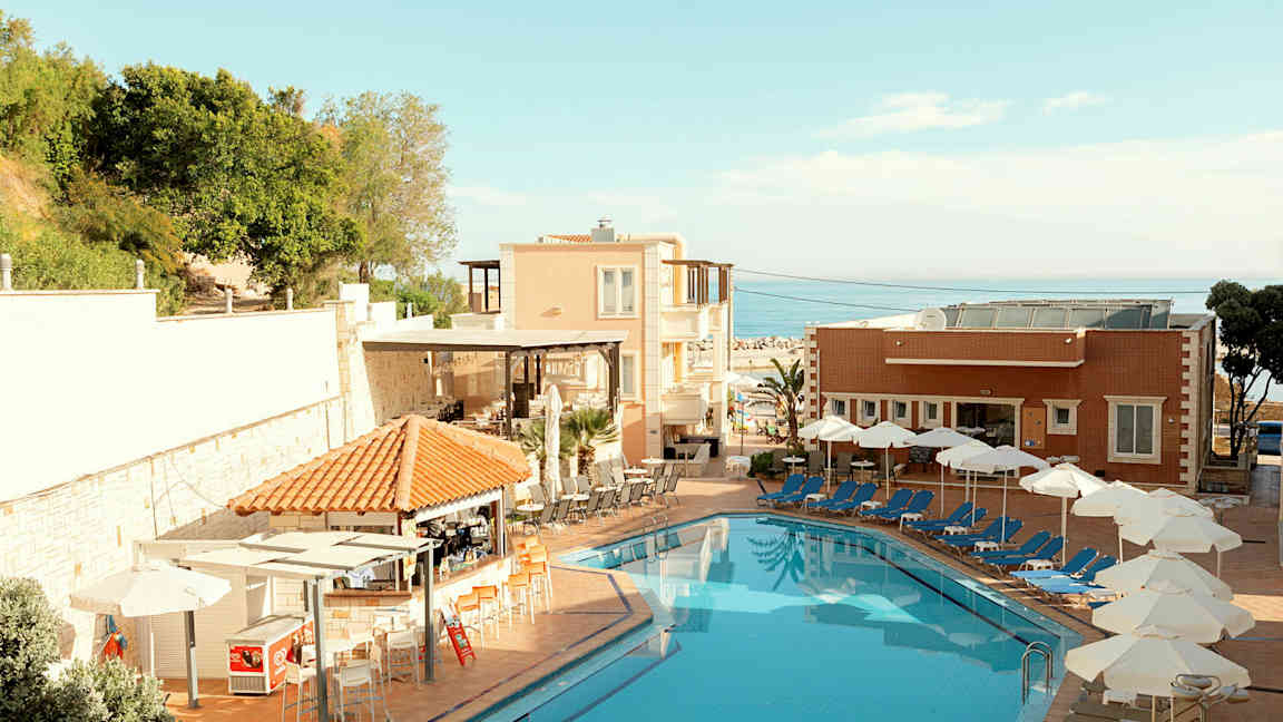 En hotellpool med solstolar och parasoller vid sidan om samt byggnader och grönska i bakgrunden. (AI-genererad text)