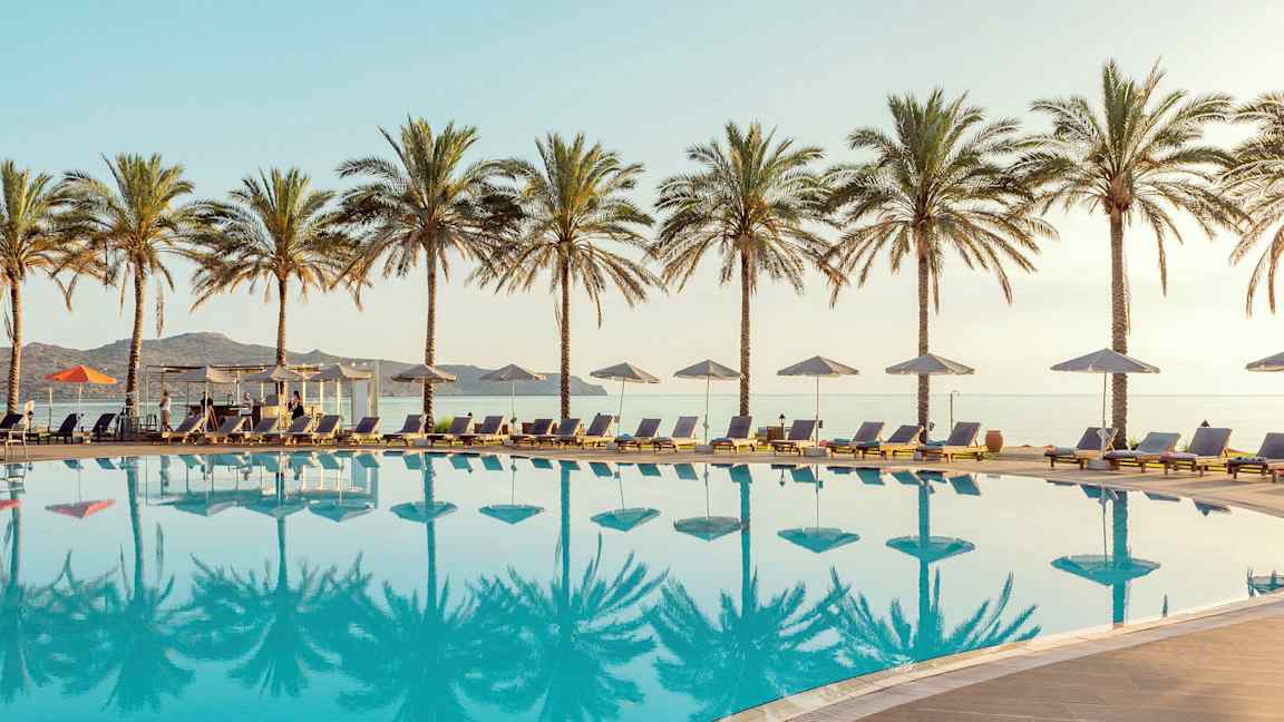 En pool med solstolar och parasoller, omgiven av palmer. Havet syns i bakgrunden. (AI-genererad text)