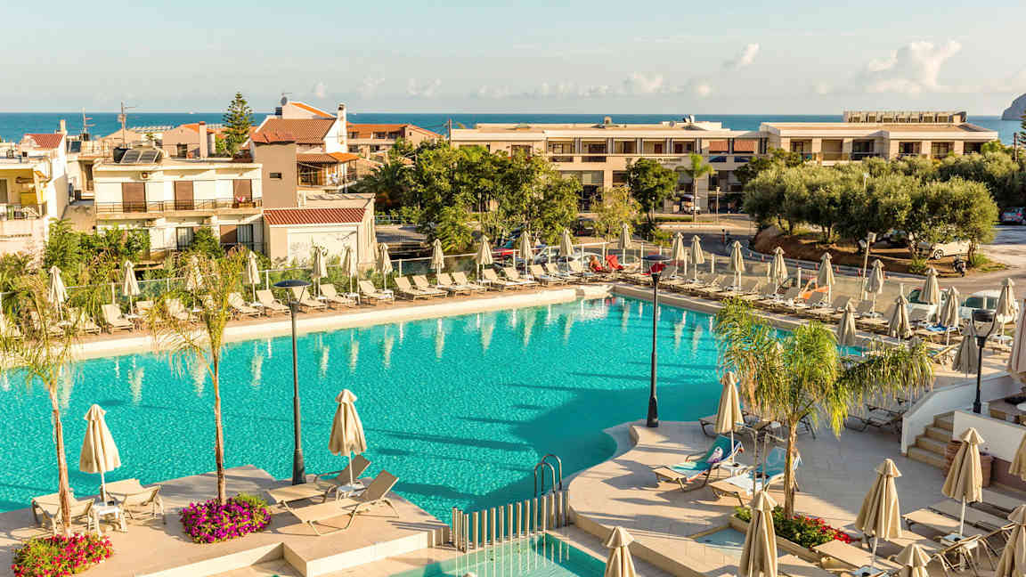 En utomhuspool omgiven av solstolar och parasoller, med byggnader och havet i bakgrunden. (AI-genererad text)