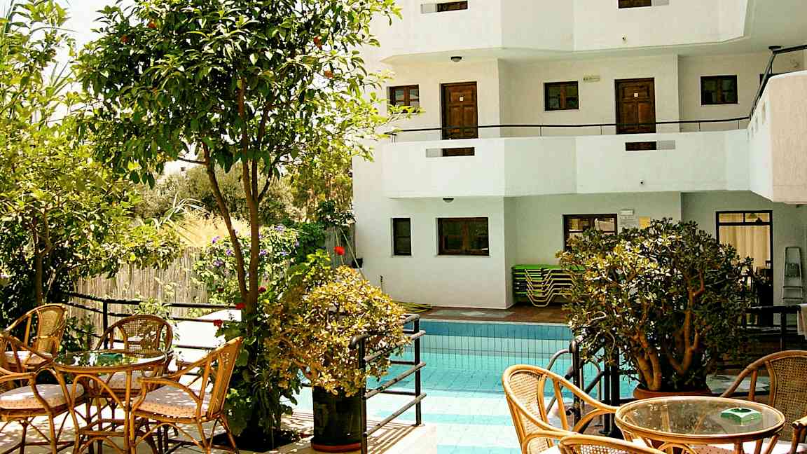 En utomhuspool omgiven av växtlighet och sittmöbler, samt en vit byggnad med balkonger i bakgrunden. (AI-genererad text)