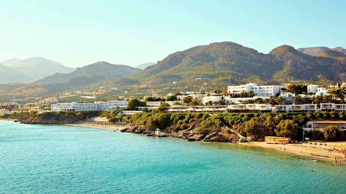 Flera byggnader nära en kust med blått hav och berg i bakgrunden. (AI-genererad text)