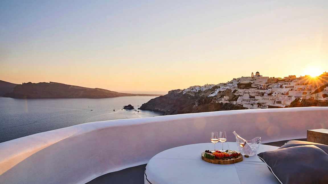 Vy över en solnedgång vid havet med vita byggnader på en klippa och en terrass med bord, mat och dryck i förgrunden. (AI-genererad text)