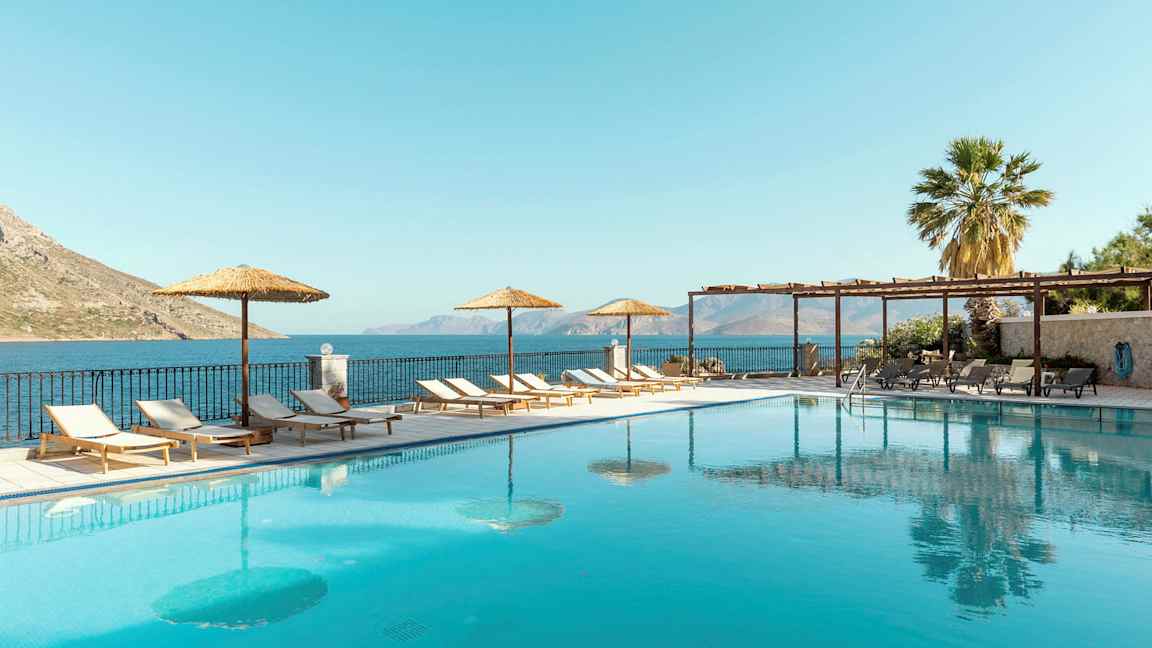 En utomhuspool med solstolar och parasoller vid sidan, samt havsutsikt i bakgrunden. (AI-genererad text)