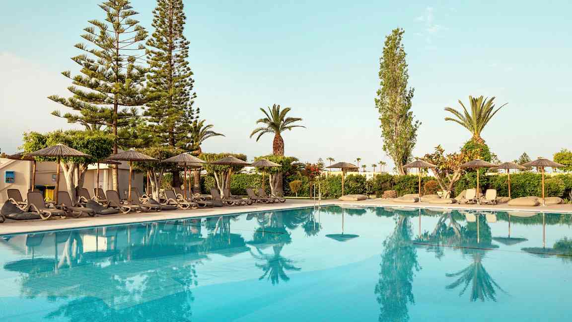 Utsikt över en pool med solstolar och parasoller vid kanten, omgiven av träd och buskar. (AI-genererad text)