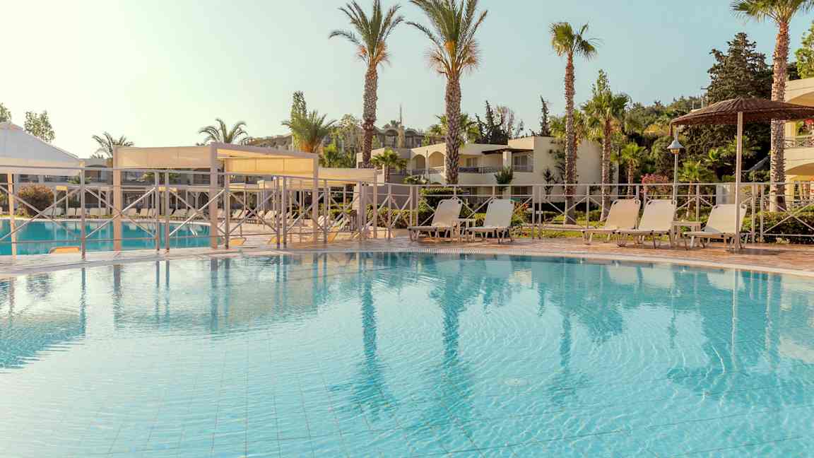 Poolområde med solstolar och parasoll, omringat av palmer och byggnader. (AI-genererad text)