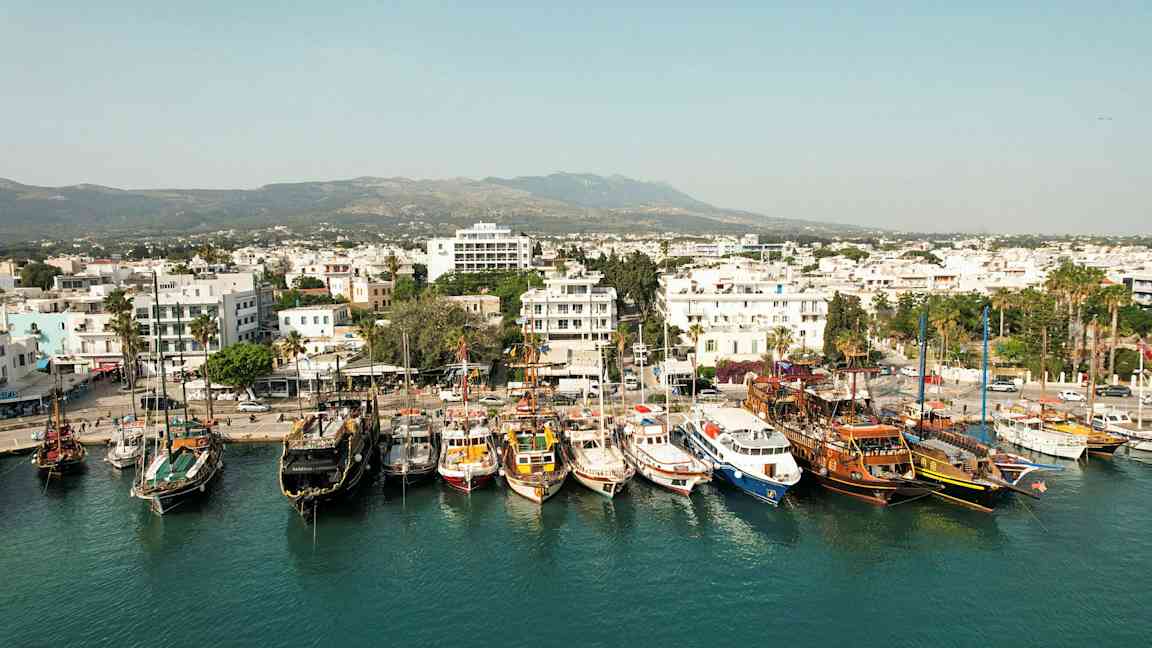 Hamnen med flera båtar förtöjda och en stad med vita byggnader i bakgrunden. (AI-genererad text)