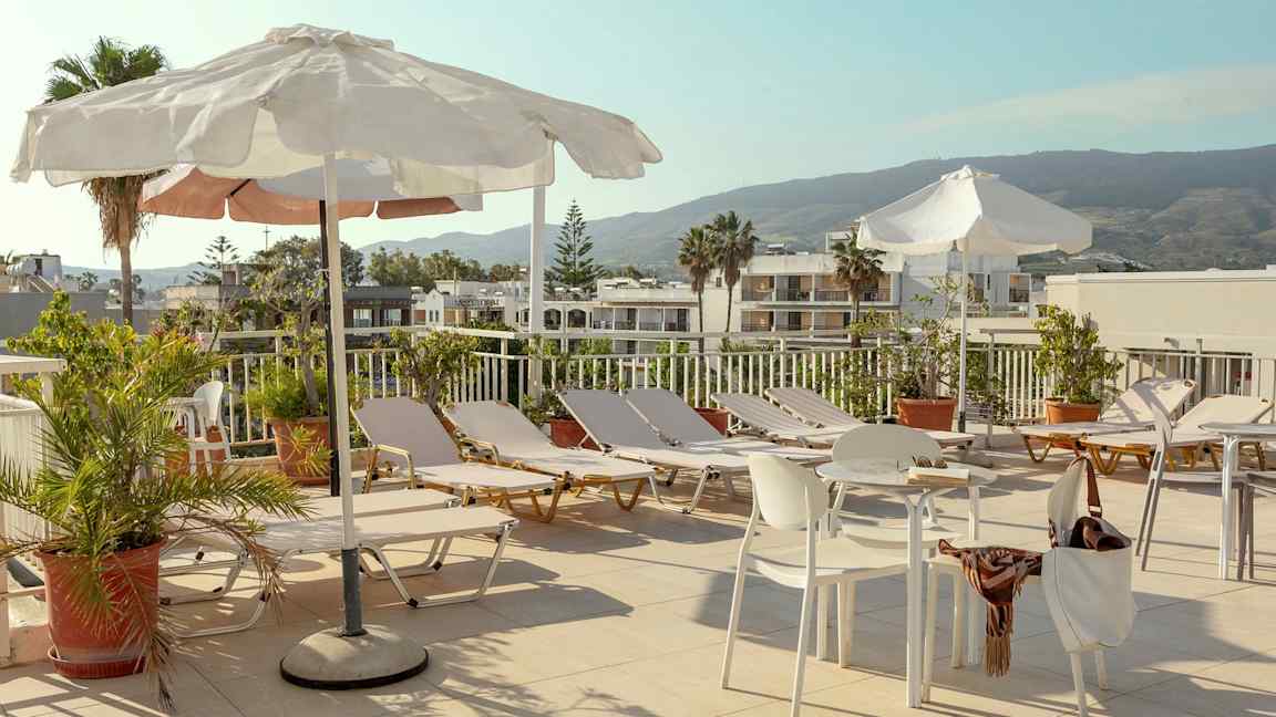 En solterrass med solstolar, parasoller och bord samt stolar omgivna av grönska och med utsikt över byggnader och berg. (AI-genererad text)