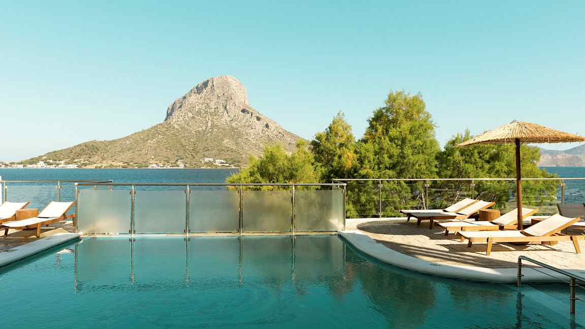 En utomhuspool med solstolar och parasoll, belägen nära havet med en kulle i bakgrunden. (AI-genererad text)