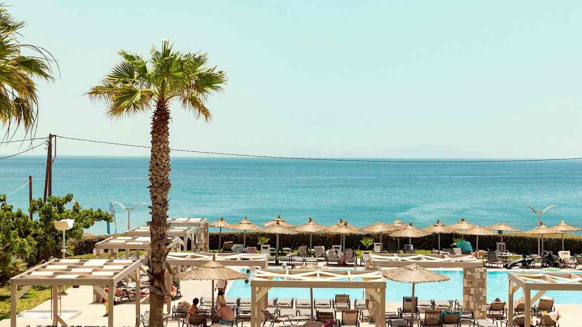 En vy över utomhuspoolområde med solstolar, parasoller, och havet i bakgrunden. (AI-genererad text)