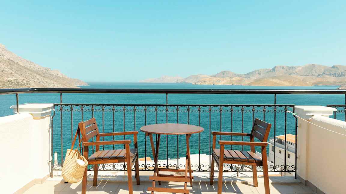 Två trästolar och ett bord på en terrass med utsikt över havet och berg i fjärran. (AI-genererad text)