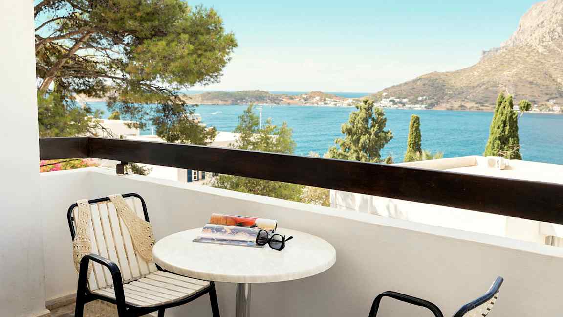 En balkong med stolar och ett bord som har en bok och solglasögon på. Utsikt över havet och en bergig bakgrund. (AI-genererad text)