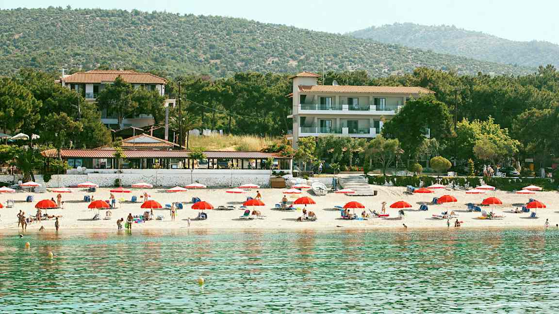 En strand med röda parasoller, solstolar och människor. Bakom stranden finns hotellbyggnader och skog. (AI-genererad text)