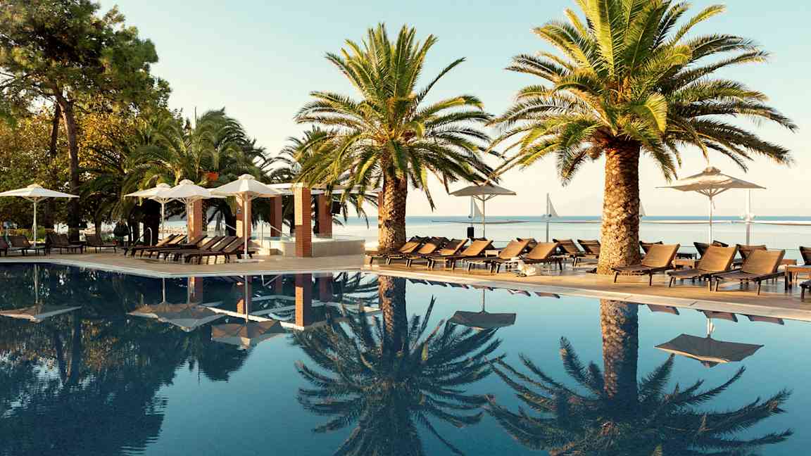 En pool med solsängar och palmer vid sidan samt utsikt mot en bro och hav. (AI-genererad text)