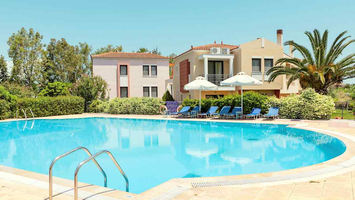 En pool omgiven av solstolar och parasoller, med byggnader och grönska i bakgrunden. (AI-genererad text)