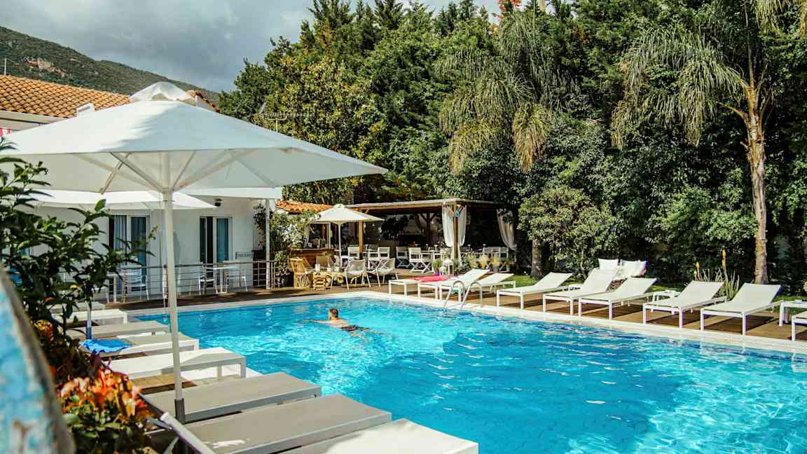 En utomhuspool omgiven av vita solstolar och parasoller nära en byggnad och grönska. (AI-genererad text)