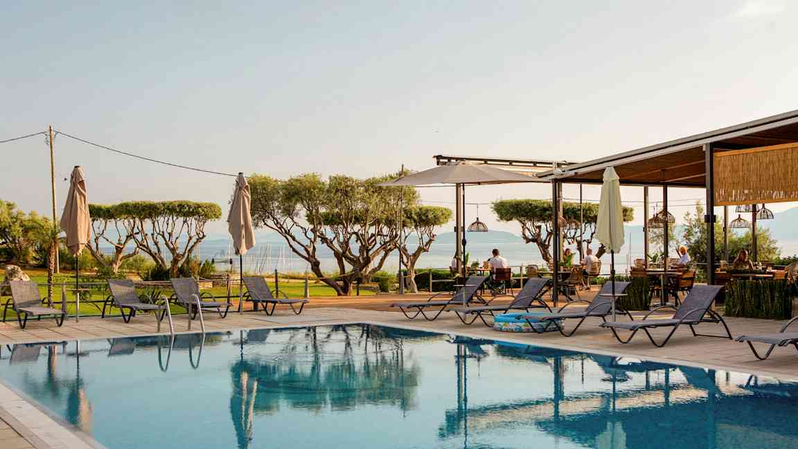 Utsikt över en pool med solstolar runtom, träd i bakgrunden och havet i fjärran. (AI-genererad text)