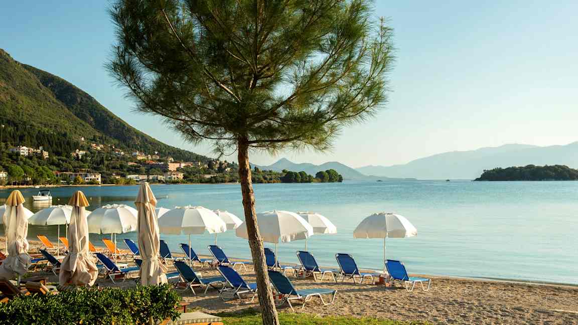 En strand med solstolar, parasoller och utsikt över havet med berg i bakgrunden. (AI-genererad text)