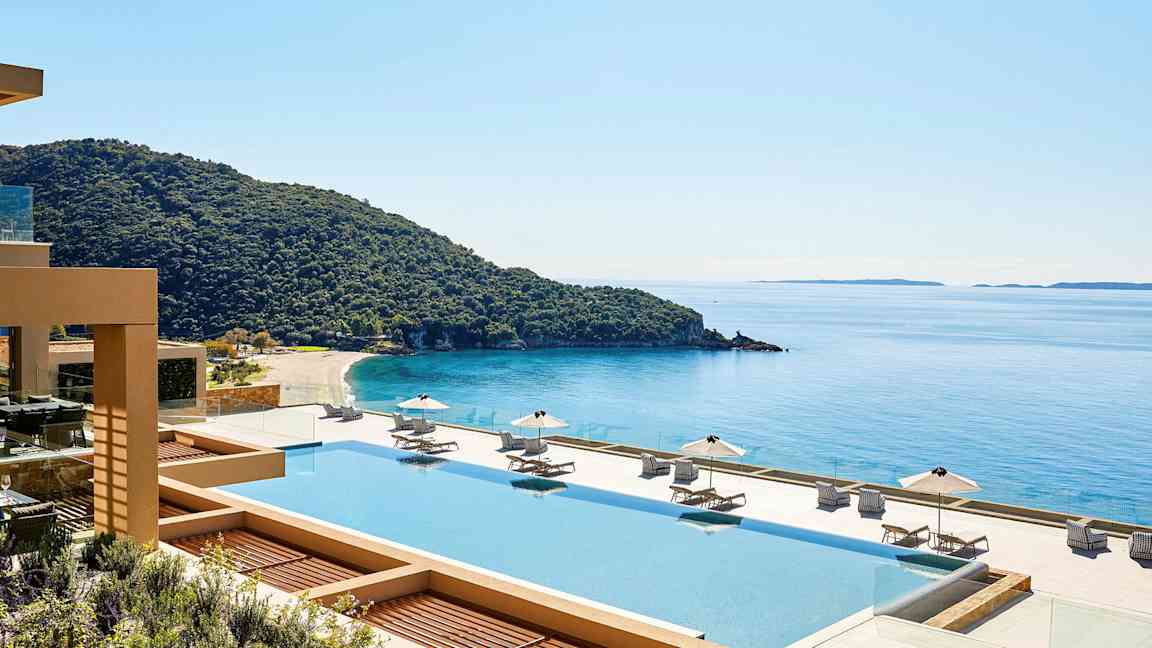 Utsikt över en pool med solstolar och parasoller, en kustlinje och hav i bakgrunden. (AI-genererad text)