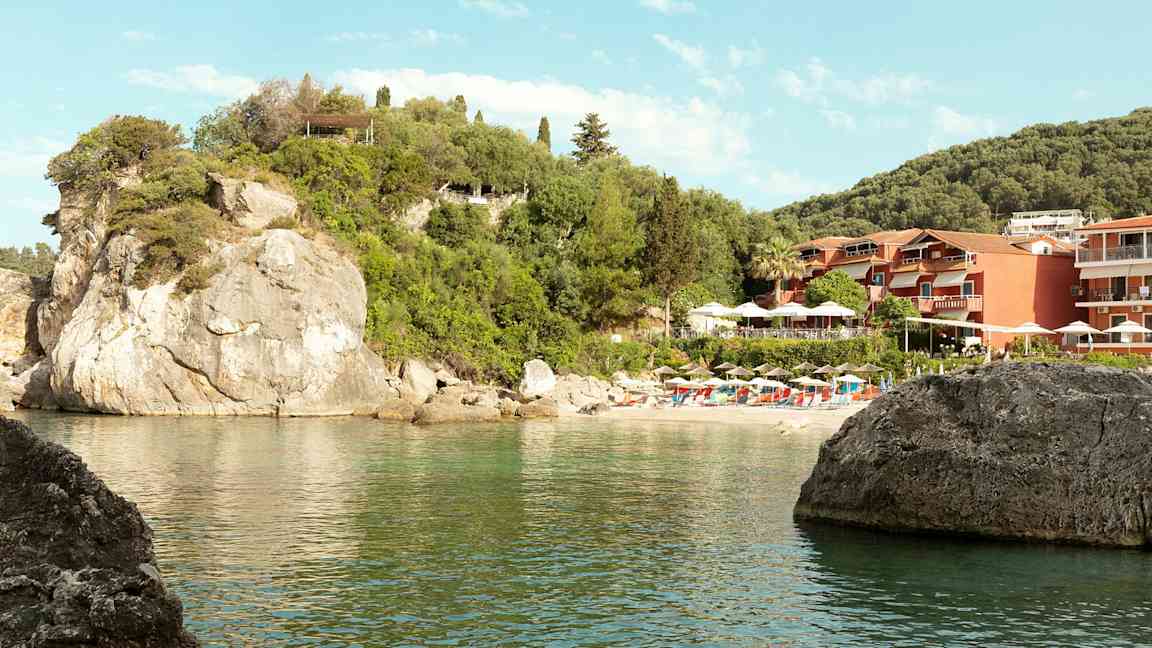 Bild av en klippstrand med parasoller och solstolar samt byggnader omgivna av grönska. (AI-genererad text)