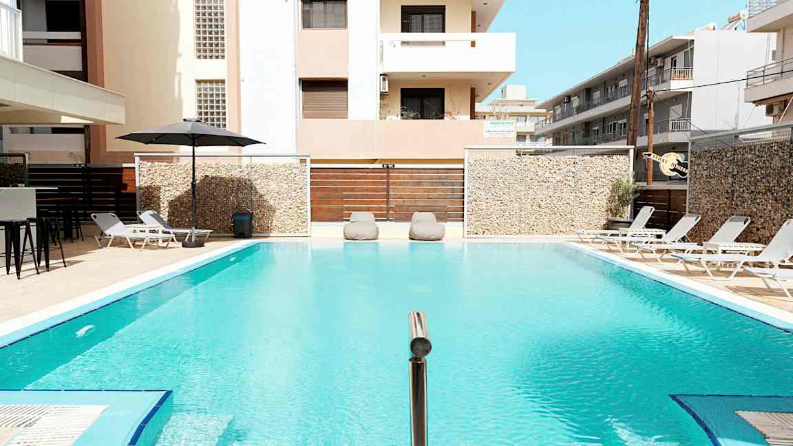 En utomhuspool omgiven av solstolar och byggnader. (AI-genererad text)