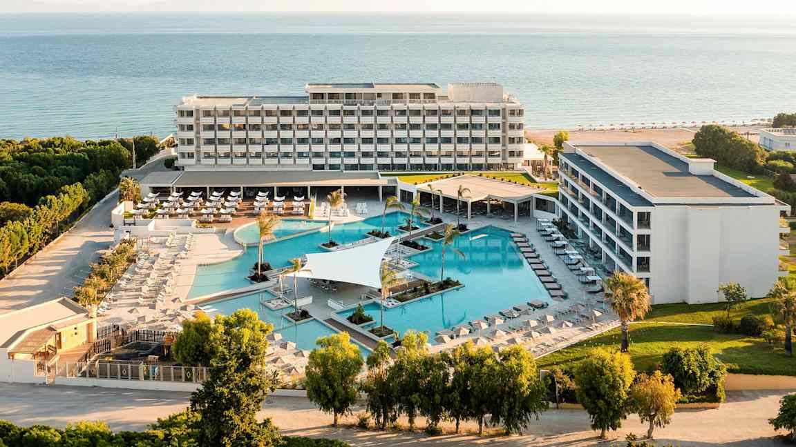 Flygfoto av ett hotellkomplex med poolområde och hav i bakgrunden. (AI-genererad text)