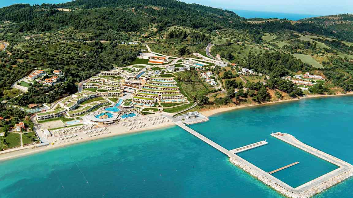 Flygbild över ett semesterområde med hotell, pooler och strand med solstolar, omgivet av grönska och hav. (AI-genererad text)