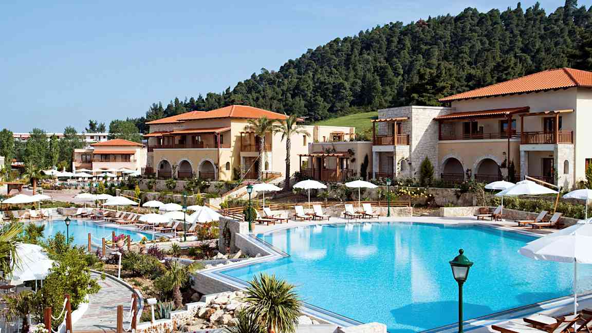 Ett hotellområde med en stor pool, omgiven av solstolar och parasoller, samt byggnader med balkonger och tegeltak. (AI-genererad text)
