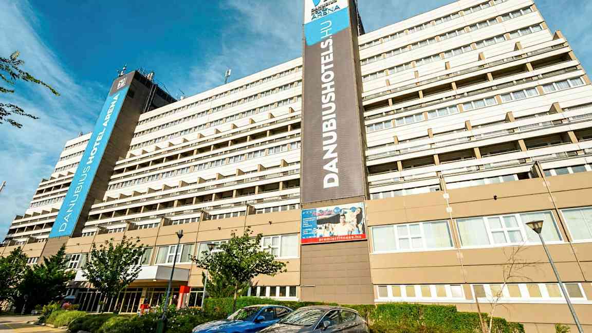 En stor hotellbyggnad med flera våningar med skylten ”Danubius Hotel Arena” synlig. Framför byggnaden finns parkerade bilar och några små träd. (AI-genererad text)
