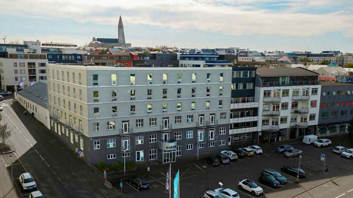 Flera moderna byggnader omger ett öppet parkeringsområde, med bilar parkerade på marken och en kyrka synlig i bakgrunden. (AI-genererad text)
