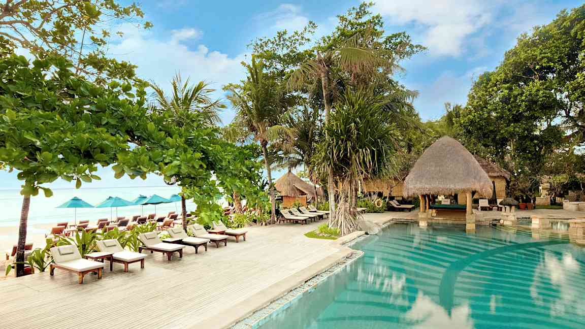 En utomhuspool omgiven av solstolar, parasoller och grönska vid havet. (AI-genererad text)