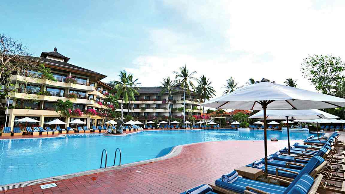 Bild av en utomhuspool omgiven av solstolar och parasoller, med ett fler vånings hotell och palmer i bakgrunden. (AI-genererad text)