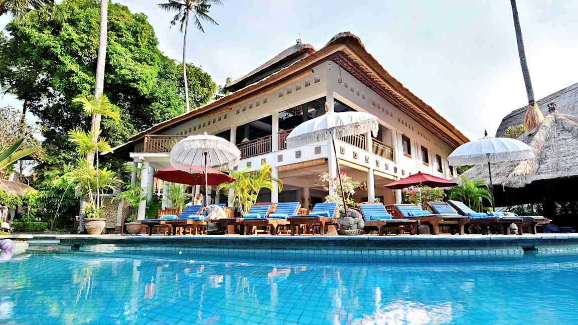 En stor villa med halmtak och en pool framför, omgiven av solstolar och parasoller. (AI-genererad text)