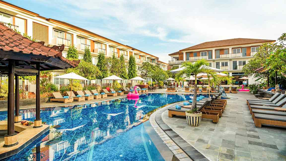 En hotellpool med solsängar och parasoller runt omkring, samt ett par byggnader i bakgrunden. (AI-genererad text)