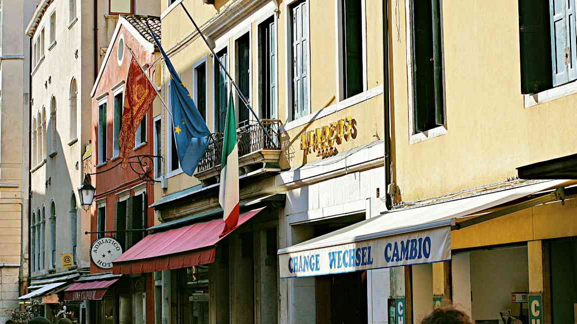 Fasad av byggnader med en italiensk flagga, europeisk flagga och skyltar som anger hotell och växling. (AI-genererad text)
