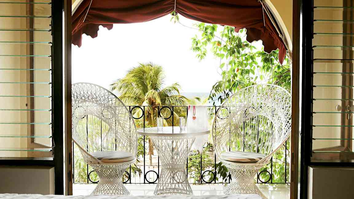 Utsikt från ett rum mot en terrass med två vita stolar och ett bord med glas, omgivna av grönska och palmer. (AI-genererad text)