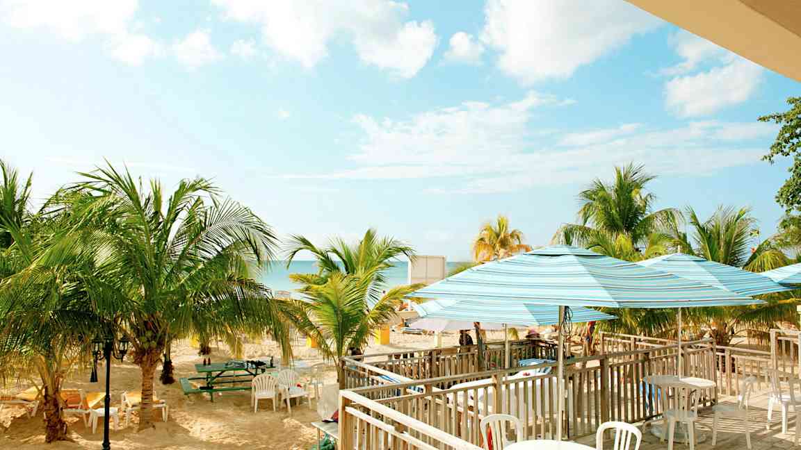 Bild av en strand med palmer, bord och stolar samt parasoll. Himlen är klar med några moln. (AI-genererad text)