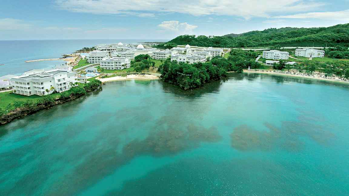 Bild av en kust med klart vatten och vita byggnader omgivna av grönska. (AI-genererad text)