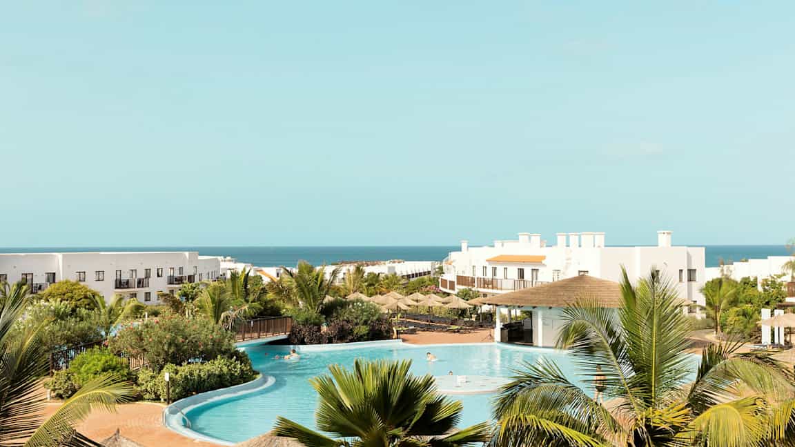 Hotellanläggning med pool, grönska och hav i bakgrunden. (AI-genererad text)