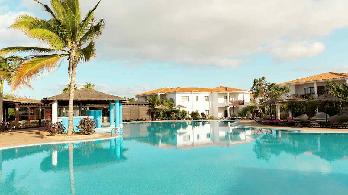 En tropisk poolmiljö med palmer, parasoller och omgivande byggnader. (AI-genererad text)