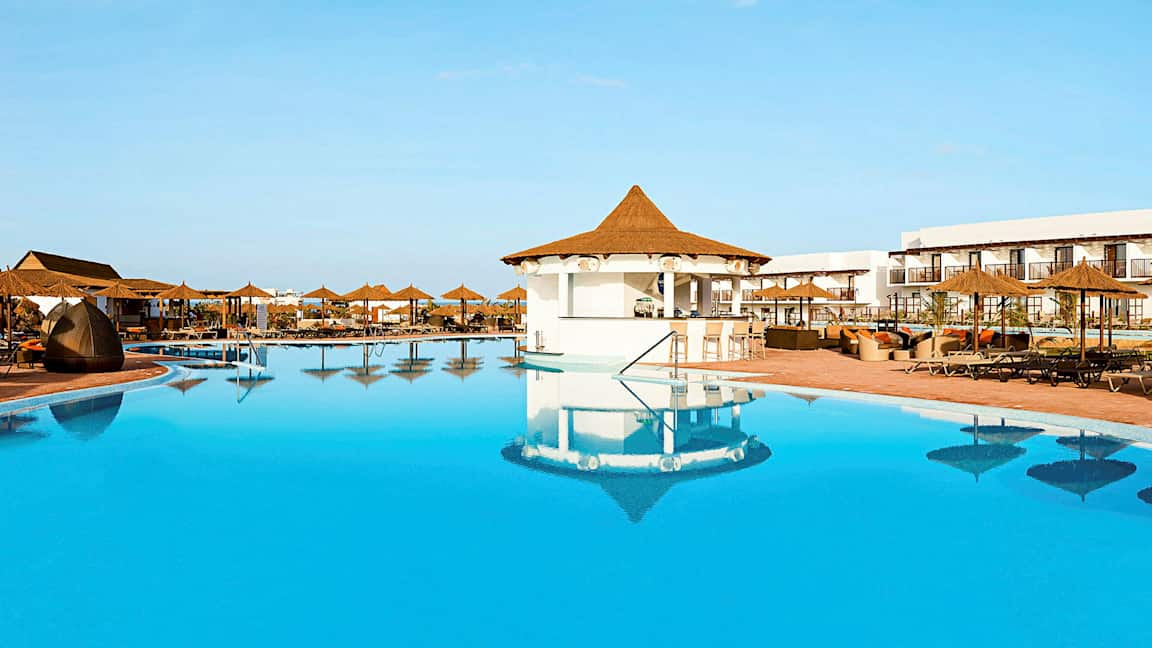 En stor pool med parasoll och sittplatser vid ett hotellområde. (AI-genererad text)