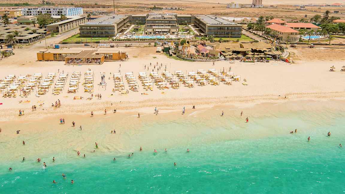 Flygfoto av en strand med turkost vatten, solstolar och parasoller, samt bakom en hotellbyggnad. (AI-genererad text)