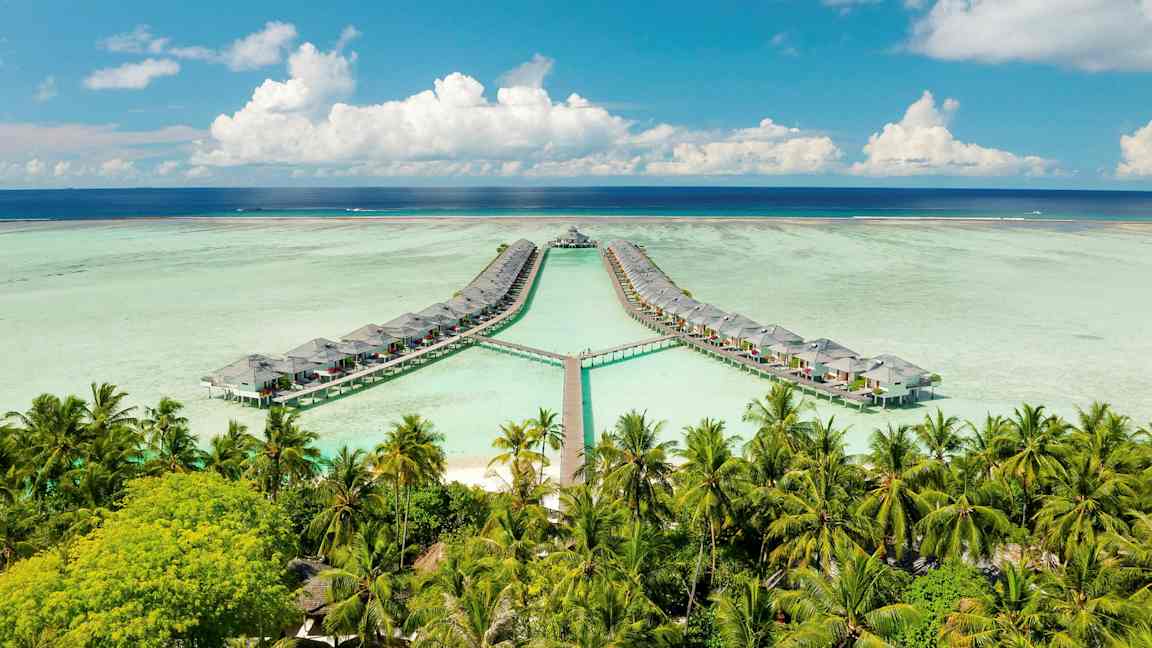 Bild av övervattenbungalower anslutna med bryggor över en lagun, omgivna av palmer och omgiven av blå himmel och hav. (AI-genererad text)