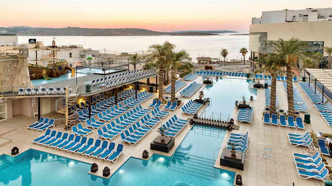 Stor poolområde med solstolar, palmer och utsikt över havet vid solnedgång. (AI-genererad text)