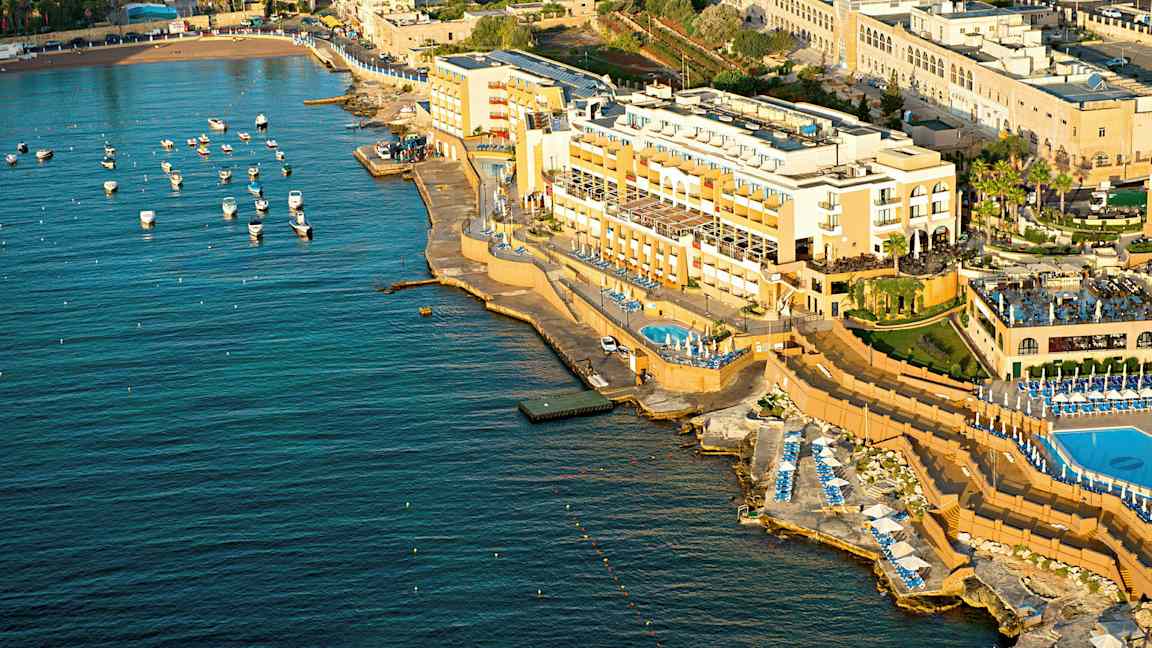 Luftvy av ett kusthotell med pooler och solstolar vid havet. Små båtar ligger förtöjda i vattnet. (AI-genererad text)