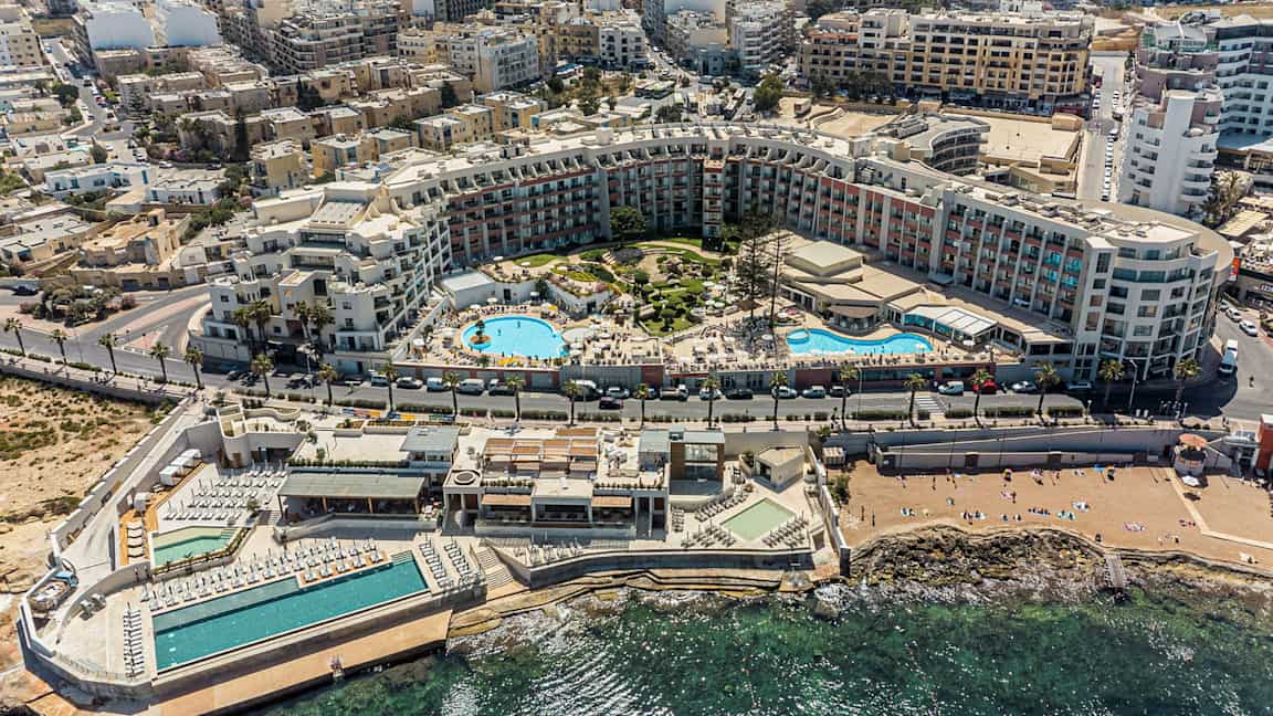 Bilden visar en omfattande semesterresort med pooler, solområden och omgivande byggnader nära kusten. (AI-genererad text)