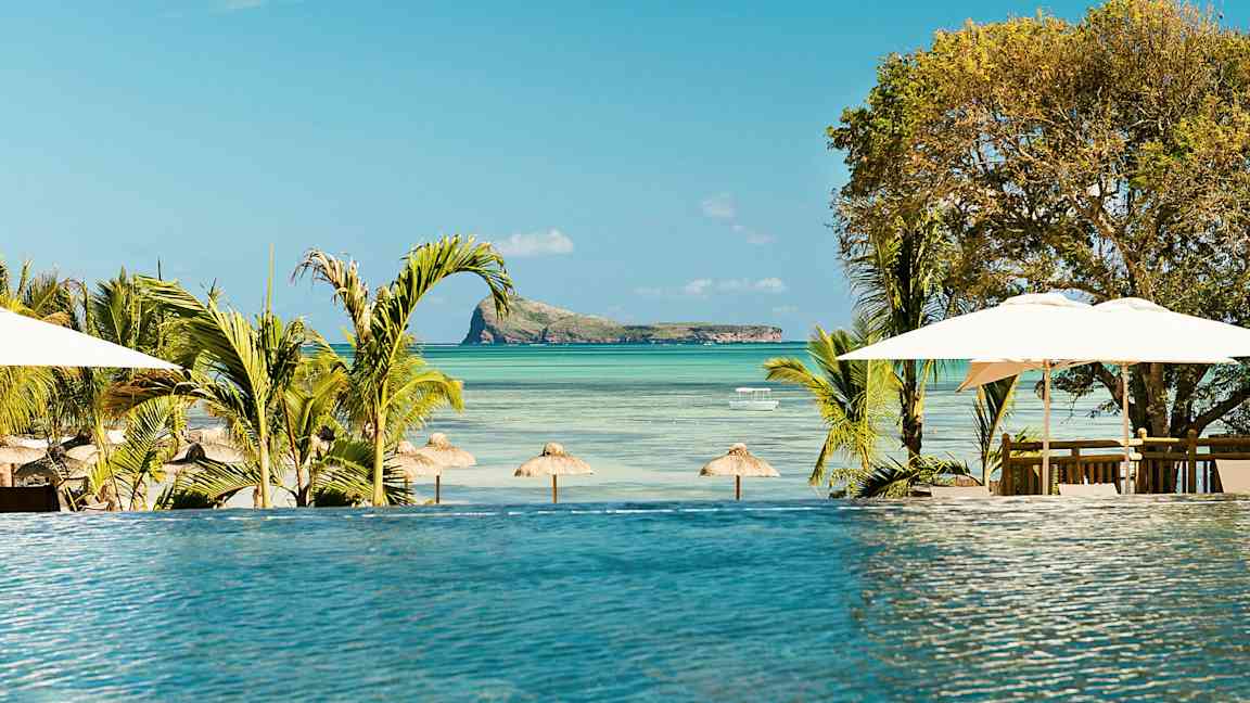 En infinitypool med utsikt över en strand med parasoller och en liten ö i havet. (AI-genererad text)