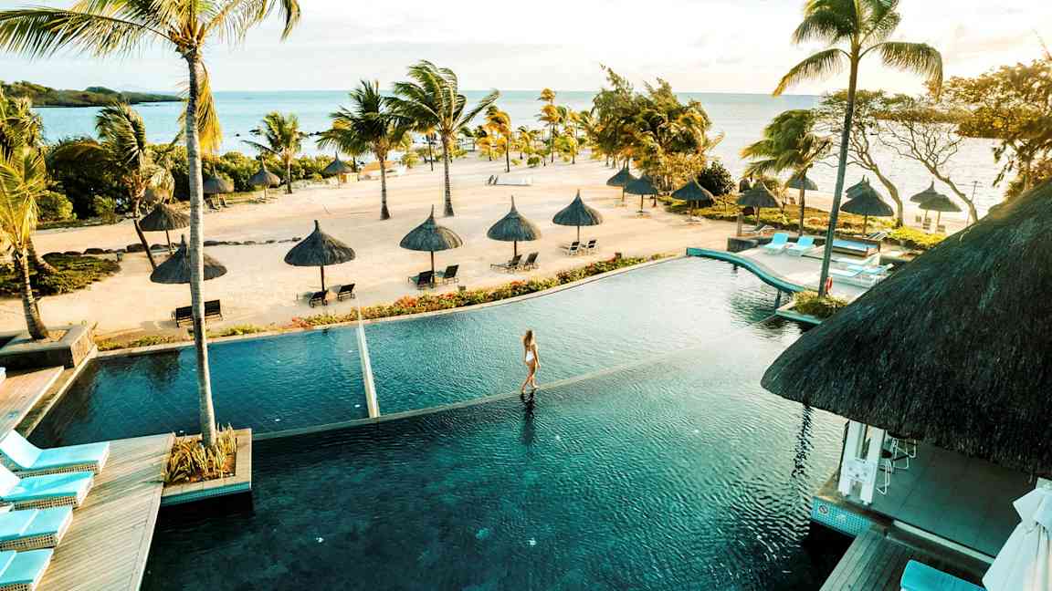 En tropisk strand med palmer och parasoller vid havet, en infinitypool i förgrunden. (AI-genererad text)