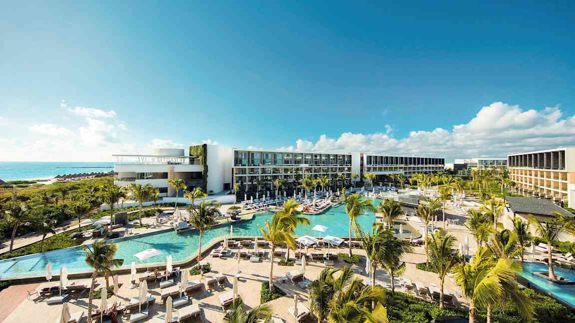 Ett modernt hotellkomplex med flera pooler, frodig vegetation och palmer, beläget vid kusten med utsikt över havet under en klarblå himmel. (AI-genererad text)