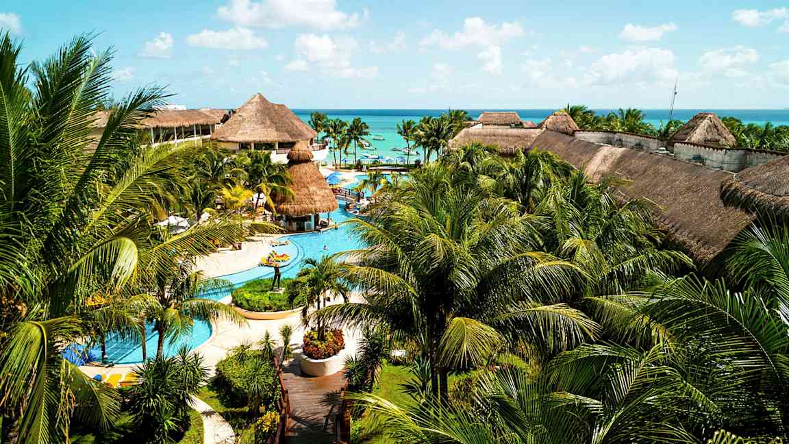 En stor resort med pooler, palmer och byggnader med halmtak, belägen nära havet under en blå himmel. (AI-genererad text)