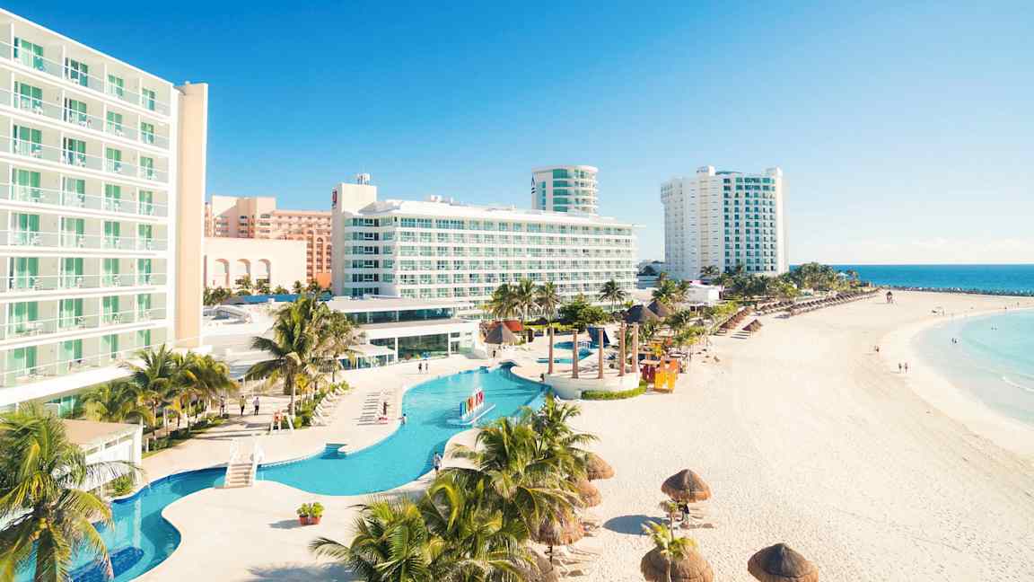 Ett hotellkomplex vid en strand med pooler i förgrunden och havsvy i bakgrunden. (AI-genererad text)